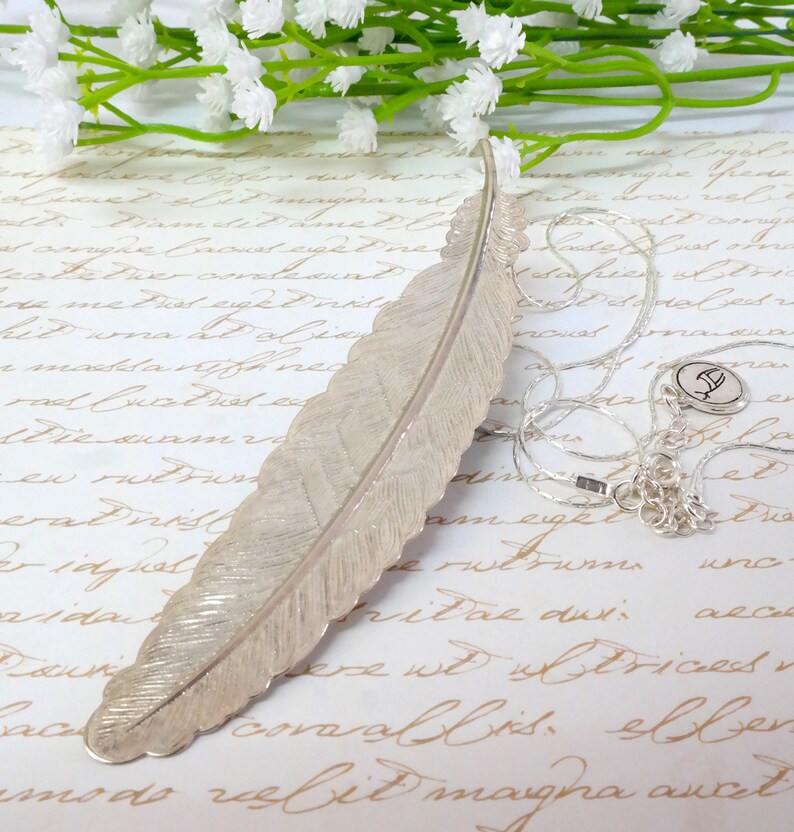 May include: A silver feather bookmark with a delicate, textured surface. The bookmark is lying on a piece of paper with handwritten text.