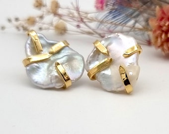 May include: A pair of white baroque pearl earrings with gold accents. The pearls are irregular in shape and have a natural luster. The gold accents are in the shape of thin, curved lines that intersect each other.