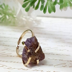 May include: A gold ring with a purple amethyst stone set in a cage-like design. The ring is on a white surface with writing.