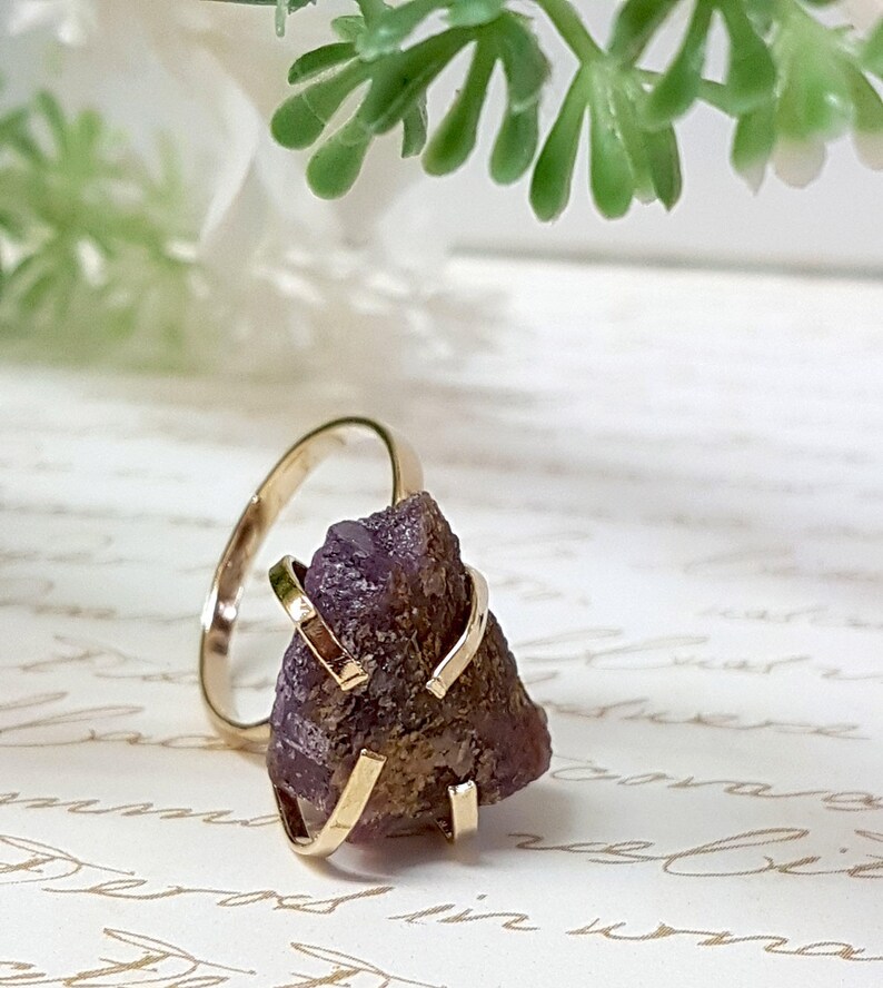 May include: A gold ring with a purple amethyst stone set in a cage-like design. The ring is on a white surface with writing.