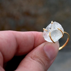 May include: A gold ring featuring a rough-cut, light blue gemstone with a small, clear accent stone. The ring's band and prongs are gold-colored, creating a contrast with the pale blue stone. The ring is held in a hand.