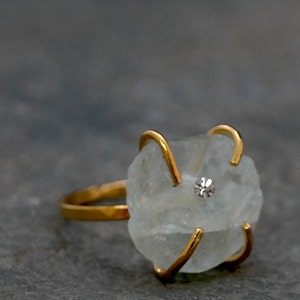 May include: A gold-toned ring featuring a light blue, irregularly shaped stone. The stone is secured by four gold prongs and has a small, round, clear gem set in the center. The ring is set against a gray background.