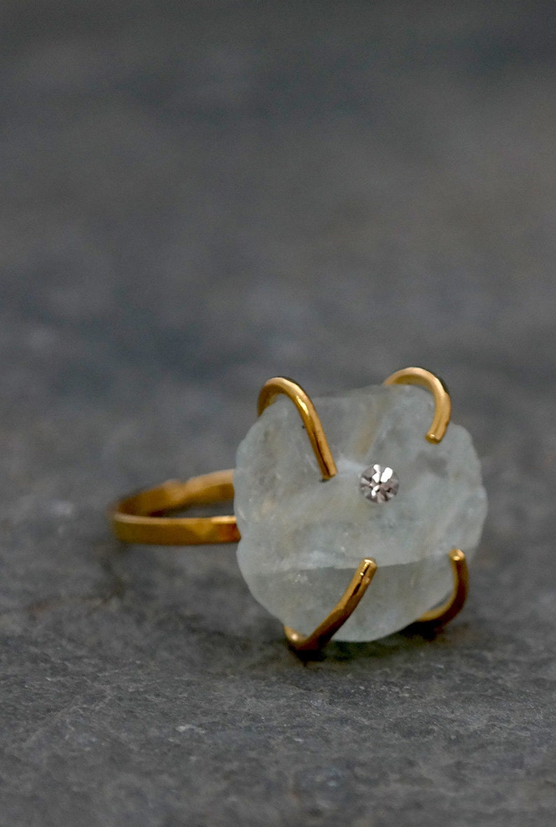 May include: A gold-toned ring featuring a light blue, irregularly shaped stone. The stone is secured by four gold prongs and has a small, round, clear gem set in the center. The ring is set against a gray background.