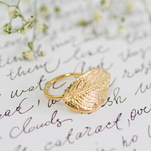 Puede incluir: Un anillo de hoja de oro con un dise&ntilde;o de hoja detallado. El anillo est&aacute; sobre una superficie blanca con texto escrito a mano.