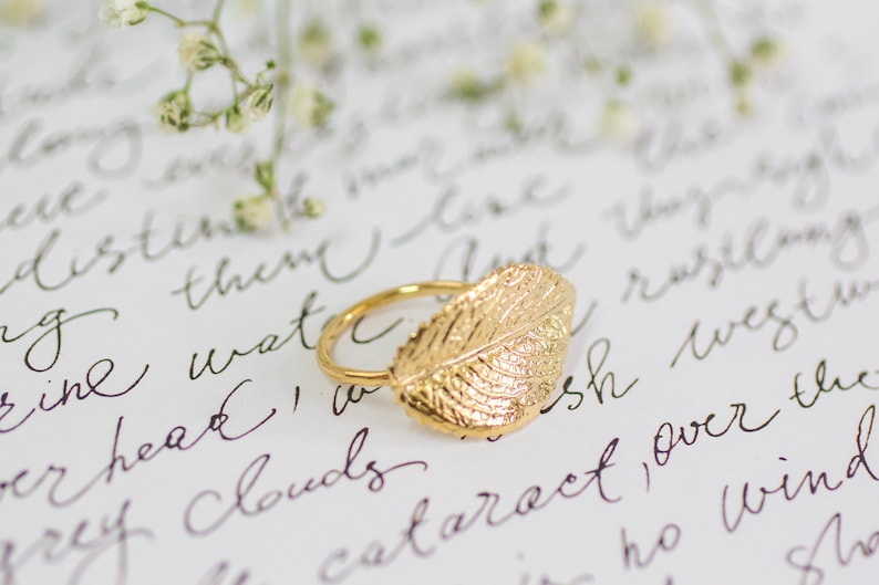 Puede incluir: Un anillo de hoja de oro con un dise&ntilde;o de hoja detallado. El anillo est&aacute; sobre una superficie blanca con texto escrito a mano.