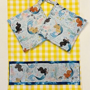 Puede incluir: Un juego de cocina con un paño de cocina a cuadros amarillos y blancos con un borde azul y dos agarraderas cuadradas a juego. Las agarraderas y el borde de la toalla tienen un fondo azul claro con ilustraciones de sirenas.