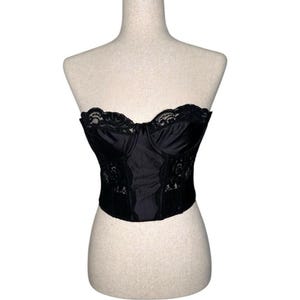 May include: Black bustier top with lace trim and a sweetheart neckline. The corset-style top is displayed on a mannequin. The bustier features a satin-like fabric and lace detailing along the edges and center.