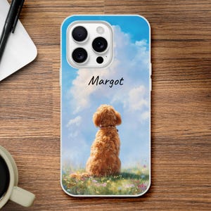 Peut inclure: Coque de téléphone avec un dessin de chien golden doodle assis dans un champ de fleurs, sur fond de ciel bleu nuageux. Le nom "Margot" est écrit en script. Le boîtier est blanc avec un bord bleu.