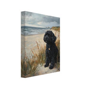 Puede incluir: Impresión en lienzo de un perro negro sentado en una playa de arena, con el mar y un cielo nublado al fondo. El perro lleva una chapa dorada. Hierba alta en primer plano.