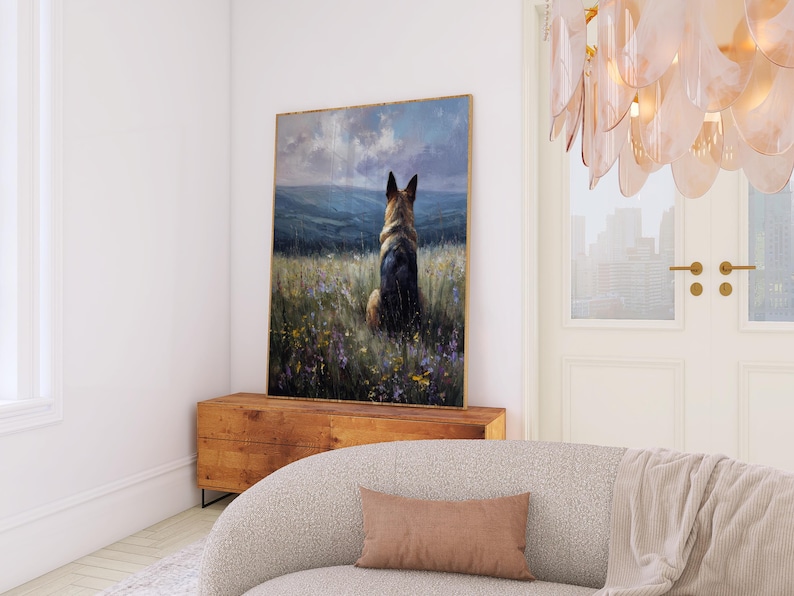 May include: A framed artwork depicts a German Shepherd dog sitting in a field of wildflowers, facing away from the viewer. The painting has a gold frame and is displayed on a wooden cabinet. The scene is set against a backdrop of rolling hills and a cloudy sky.