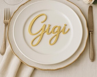 May include: A gold-colored, cursive "Gigi" nameplate rests on a white plate with a gold rim. The plate is set on a larger white plate with a gold rim, with a fork, knife, and napkin visible. A rose and greenery are in the background.