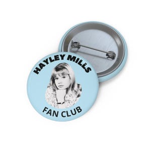 May include: A light blue button with the text "HAYLEY MILLS FAN CLUB" in black. The button features a black and white image of Hayley Mills. The button is round and has a metal pin back.
