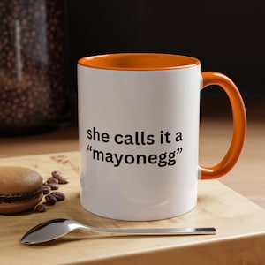 Peut inclure: Mug en céramique blanche avec intérieur et anse orange. Le mug porte l'inscription noire "she calls it a 'mayonegg'". Une cuillère, un macaron et des grains de café sont sur une surface en bois.