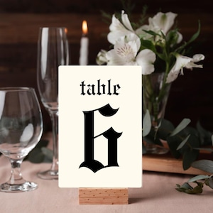 Op de afbeelding: Een tafelnummerkaart met het woord "tafel" en het nummer "6" in een zwart gotisch lettertype. De kaart is crèmekleurig en staat in een houten houder. De achtergrond omvat wijnglazen, bloemen en kaarsen.