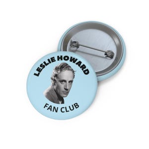 May include: A light blue Leslie Howard Fan Club button with a black and white portrait of Leslie Howard. The text "LESLIE HOWARD" arcs around the top, and "FAN CLUB" is at the bottom. The button is round and has a metal pin back.