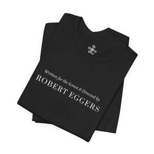 Könnte beinhalten: Schwarzes T-Shirt mit weißem Text, der lautet: "Written for the Screen & Directed by ROBERT EGGERS". Das Shirt hat auch den Text "The Gloaming Press" in der Nähe des Kragens. Das T-Shirt ist gefaltet.