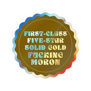 May include: A brown, scalloped-edge sticker with the words "FIRST-CLASS FIVE-STAR SOLID GOLD FUCKING MORON" in a rainbow gradient. The sticker has a dotted inner circle and a metallic sheen.