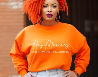 May include: An orange sweatshirt with the text "His Mercies Are New Every Morning" in white script. The sweatshirt is paired with light-wash jeans. The person in the image has orange hair and is wearing hoop earrings.