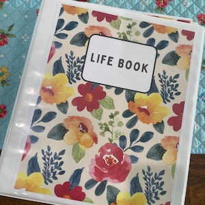 May include: A white binder with a floral design cover. The cover features a cream background with watercolor-style flowers in red, yellow, and orange, along with blue leaves. The words "LIFE BOOK" are printed on a white label.
