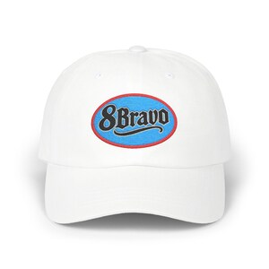 May include: White baseball cap with a curved brim. The cap features an embroidered oval patch with the text "8 Bravo" in a black, stylized font, outlined in red and blue.