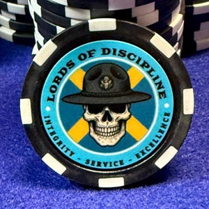May include: A poker chip with a black and white edge, featuring a skull wearing a hat. The chip has the words "LORDS OF DISCIPLINE" and "INTEGRITY - SERVICE - EXCELLENCE" around the edge.