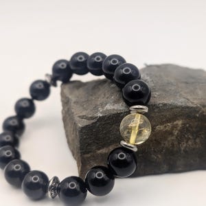 May include: A black beaded bracelet with a single yellow-tinted bead. The bracelet is made of round, glossy black beads and is accented with silver-colored metal spacers. The bracelet is resting on a gray stone.
