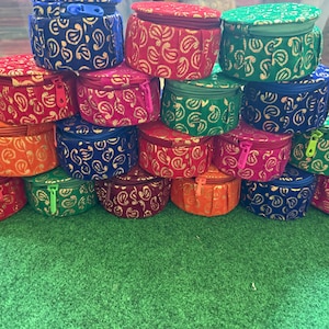 May include: A collection of small, round, fabric-covered boxes in various colors, including red, blue, green, and orange. Each box features a gold paisley pattern and a matching zipper closure. The boxes are stacked on a green surface.
