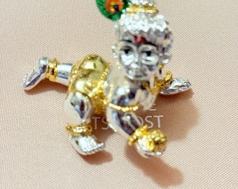 May include: A silver and gold figurine of a crawling baby, adorned with a green and gold peacock feather. The baby has a red dot on its forehead and wears gold jewelry. The figurine is likely a religious or cultural item.