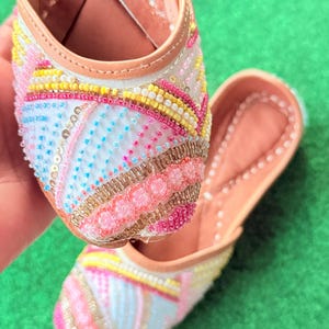 May include: Close-up of a pair of embellished shoes. The shoes feature a light pink, white, yellow, and blue beaded design. The soles are a light brown color, and the shoes are set against a green background.