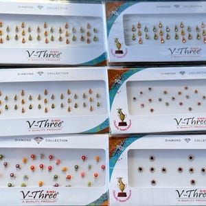 May include: Assortment of V-Three brand bindi stickers in various colors and designs. The stickers are displayed in clear plastic packaging with the brand name and "Diamond Collection" printed on the boxes.