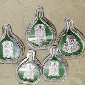 May include: Five clear, leaf-shaped display cases with silver religious icons on a green background. The icons depict various deities and figures. The cases are arranged on a neutral-toned surface.