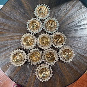 May include: Arrangement of eleven gold-colored, sunburst-shaped candle holders on a dark wooden table. Each holder has a central cup and a decorative, scalloped edge with a light blue accent. The holders are arranged in a triangular pattern.