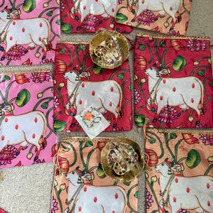 May include: Drawstring bags in shades of pink, red, and peach, each displaying a white cow illustration with red handprints. The bags are decorated with floral and leaf motifs, and some hold small, gold decorative bowls with jewelry.