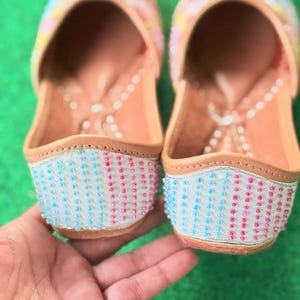 May include: A pair of colorful, beaded slip-on shoes. The uppers are decorated with pink, yellow, and blue accents. The toe and heel sections are adorned with rows of blue and pink beads. The shoes have a light brown leather sole.