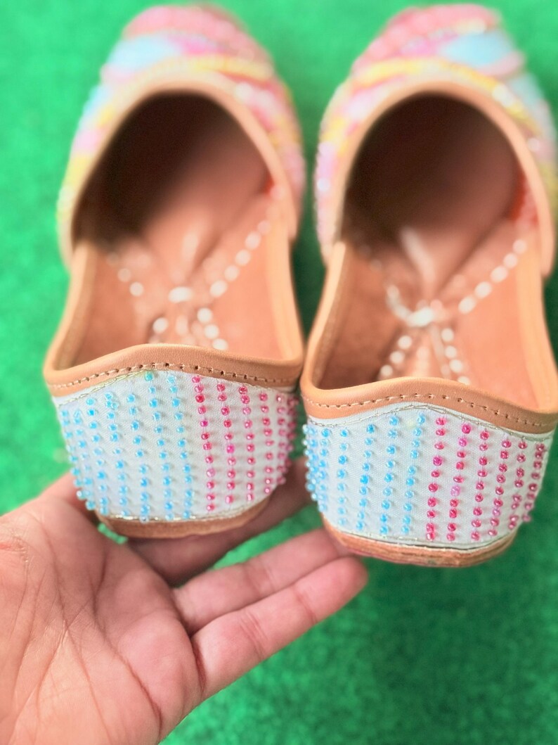May include: A pair of colorful, beaded slip-on shoes. The uppers are decorated with pink, yellow, and blue accents. The toe and heel sections are adorned with rows of blue and pink beads. The shoes have a light brown leather sole.