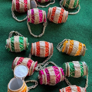 May include: A collection of colorful, cylindrical, beaded purses in shades of orange, pink, green, and red. Each purse has a beaded handle and is decorated with gold trim and a zipper closure. The purses are arranged on a green surface.