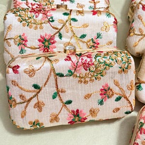 May include: Ivory-colored rectangular clutch bags with floral embroidery in pink, red, green, and gold. The bags have a gold clasp and frame. The floral design covers the entire surface of the bags.