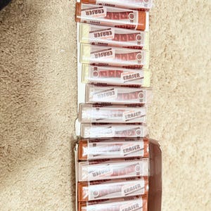May include: A box of erasers, each individually packaged in a clear plastic sleeve. The erasers are rectangular, with a brown and white color scheme. The word "ERASER" is printed on each package in a bold, red font.