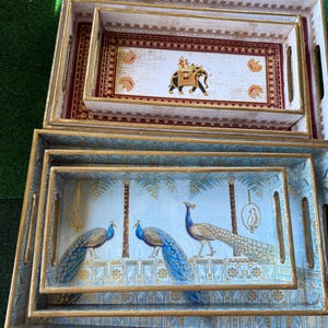 May include: Set of four decorative wooden trays. Two trays feature a design with peacocks in blue and gold. The other two trays have an elephant design in gold, red, and white. The trays have gold trim.