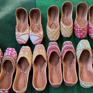 May include: A collection of colorful, embellished leather slip-on shoes. The shoes feature intricate beadwork and embroidery in various colors, including pink, gold, and blue. The soles are a light brown color, and the shoes are arranged on a green surface.