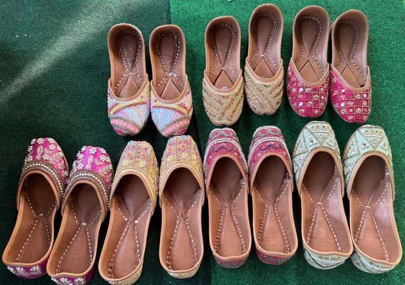 May include: A collection of colorful, embellished leather slip-on shoes. The shoes feature intricate beadwork and embroidery in various colors, including pink, gold, and blue. The soles are a light brown color, and the shoes are arranged on a green surface.
