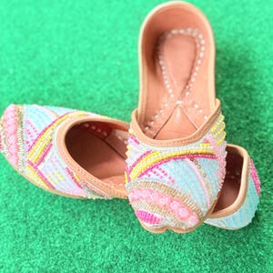 May include: A pair of colorful, beaded slip-on shoes. The shoes feature a light pink, blue, and yellow beaded design with a tan leather sole and trim. The shoes are displayed on a green surface.