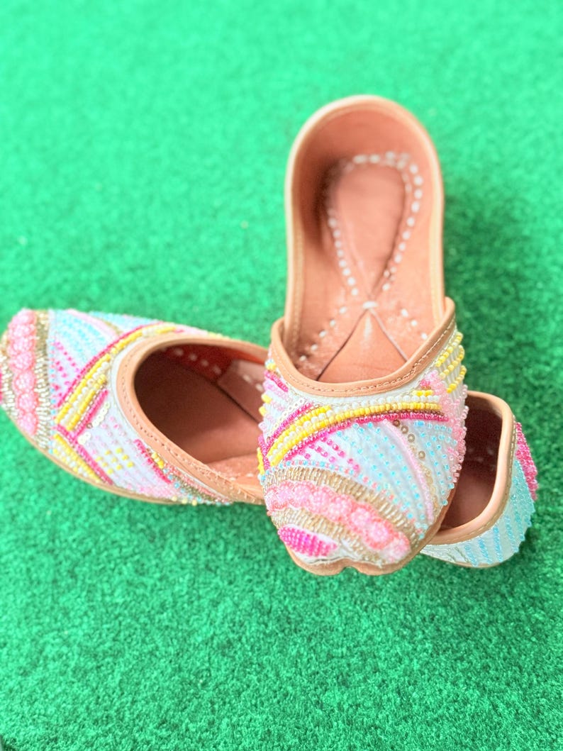May include: A pair of colorful, beaded slip-on shoes. The shoes feature a light pink, blue, and yellow beaded design with a tan leather sole and trim. The shoes are displayed on a green surface.
