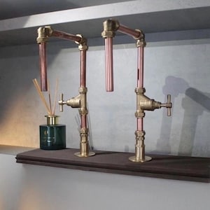 Può includere: Due elementi decorativi a forma di rubinetto in rame e ottone montati su una mensola in legno scuro. Gli elementi hanno un design industriale vintage, con tubi in rame e raccordi in ottone. Un diffusore in vetro verde con canne è a sinistra.