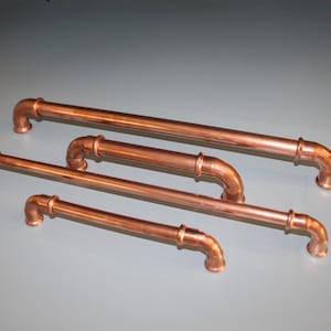 May include: Four copper-colored pipe-style cabinet pulls in varying lengths. Each pull features a straight bar with curved ends and connecting joints. The hardware is arranged on a gray surface, showcasing a rustic, industrial design.