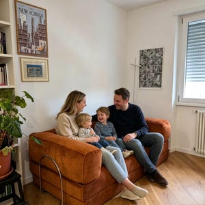 May include: A family of four sits on a burnt orange corduroy sofa in a well-lit living room. The parents are smiling at their two young children. Artwork and a bookshelf are visible in the background, creating a cozy home atmosphere.