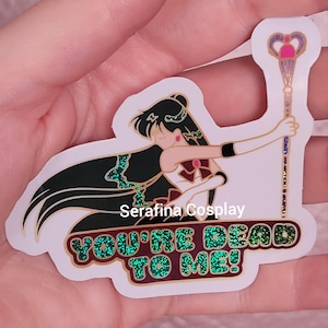 May include: A white sticker with a green, black, and red anime character holding a gold and purple staff. The character has long black hair and is wearing a white and red outfit. The sticker says "You're Dead To Me!" in green glitter letters.
