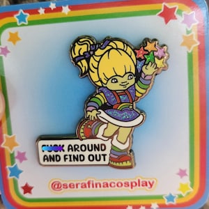 May include: A colorful enamel pin featuring a cartoon character with blonde hair and a rainbow outfit, holding star-shaped objects. The pin includes the text "FUCK AROUND AND FIND OUT" on a white rectangular banner. The pin is on a card with a rainbow and star design.
