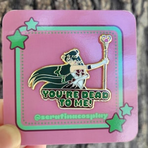 May include: A metal enamel pin featuring an anime-style character with long dark hair holding a staff, set against a pink backing card with green stars. The pin includes the text "YOU'RE DEAD TO ME!" in green glitter and "@serafinacosplay" below.