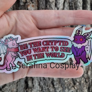 May include: A holographic sticker with a pink dragon and a purple moth. The sticker says "Be the cryptid you want to see in the world" and "Serafina Cosplay".